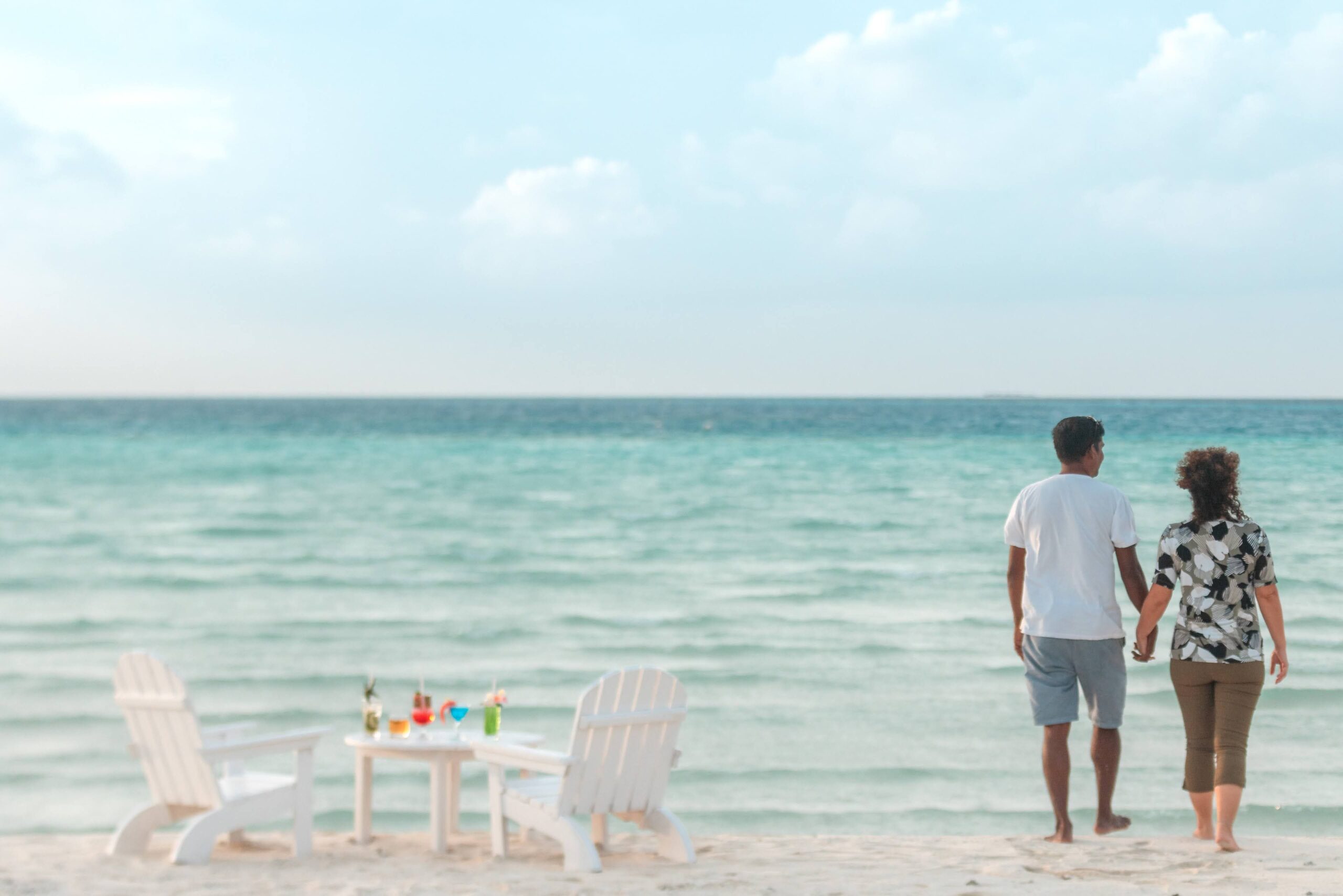 Couple enjoying a serene walk on a tropical beach with chairs and drinks.